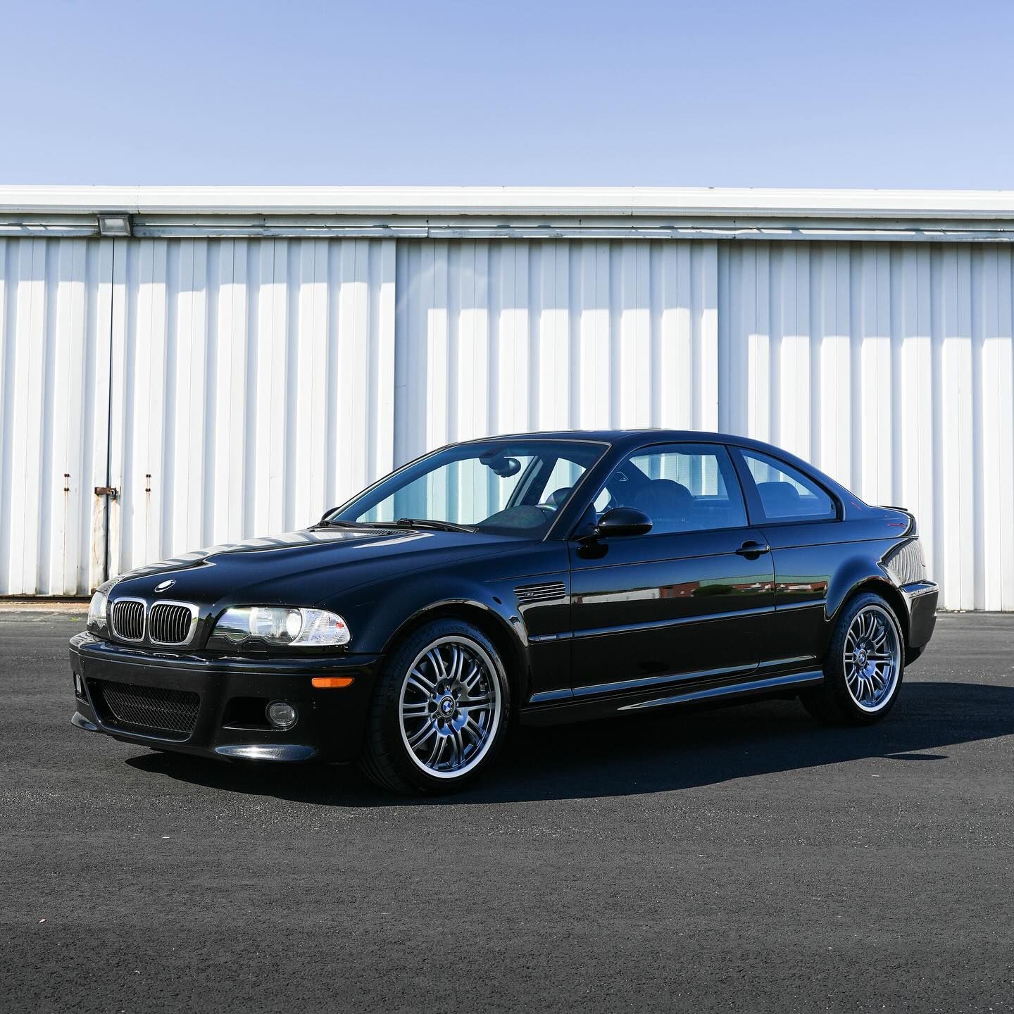 Black BMW M3 coupe parked on asphalt, in front of a white building on a sunny day.