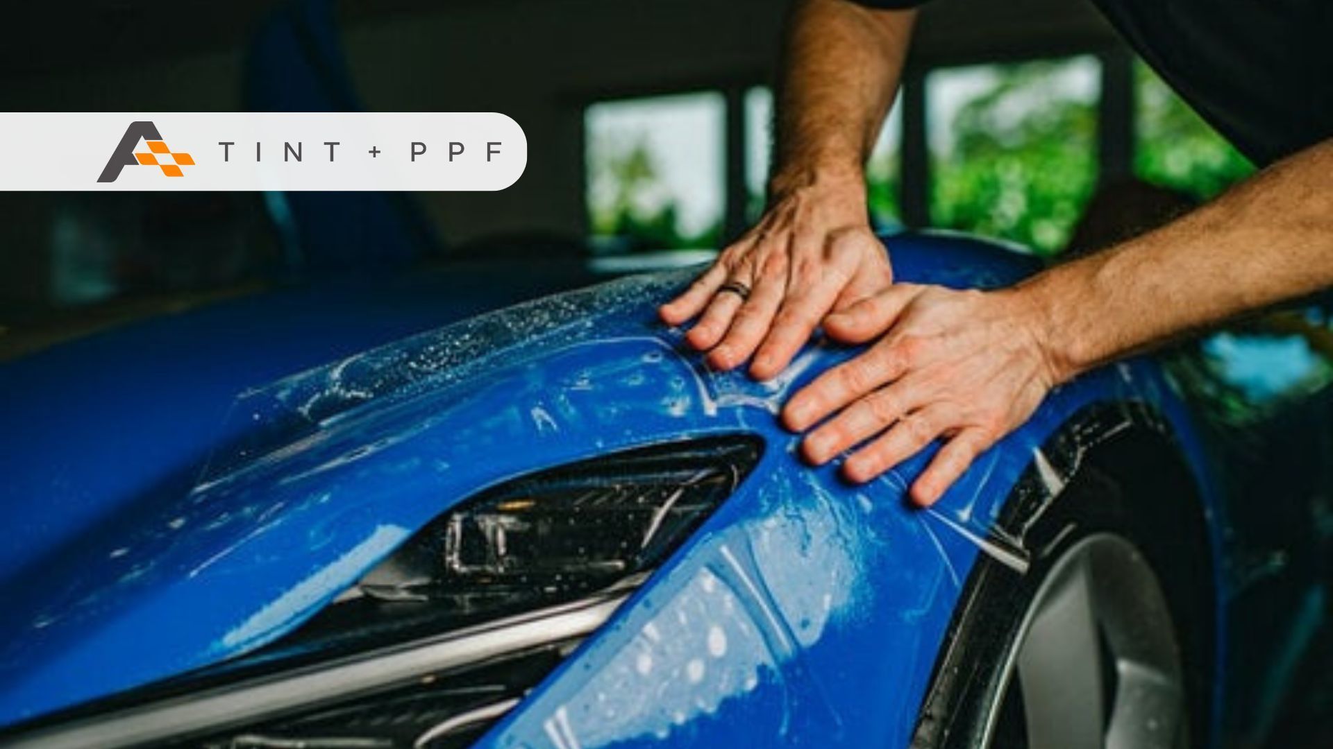 Two hands smoothing paint protection film onto the curved fender of a blue sports car