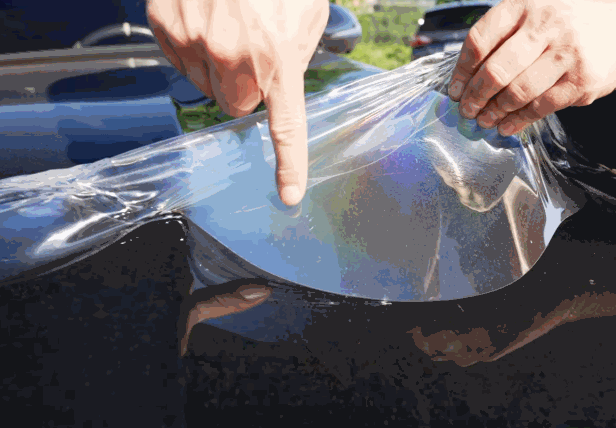 A technician peeling back a portion of paint protection film (PPF) from a black car, carefully applying it to the surface.