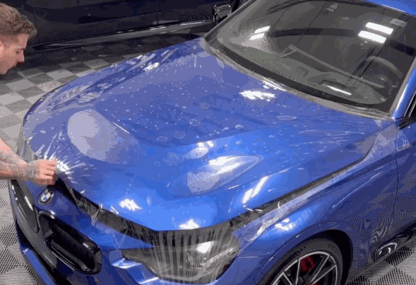 A technician applying paint protection film (PPF) to the hood of a blue BMW, ensuring the film is smooth and bubble-free.