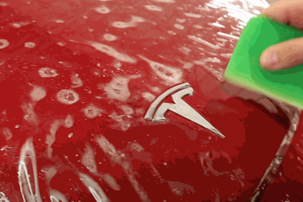 A close-up of a person using a squeegee to smooth out the paint protection film (PPF) over the Tesla logo on a red car.