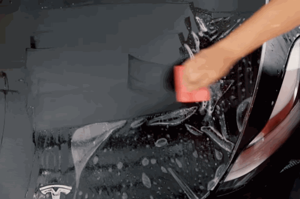 A person smoothing out the paint protection film (PPF) over the front of a black Tesla using a red squeegee.