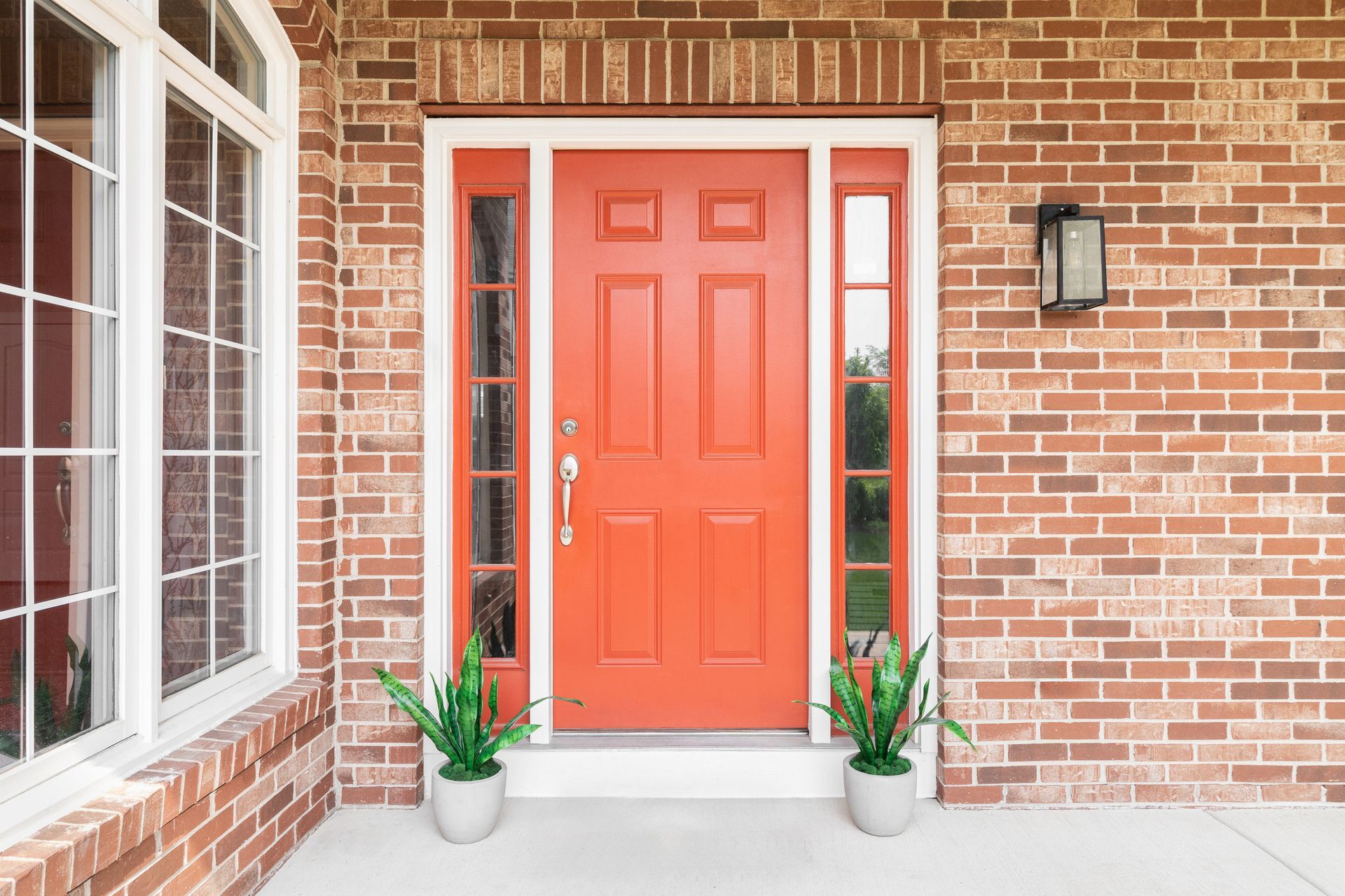 Entry door replacement featuring orange door with glass sidelights on brick home and potted plants.