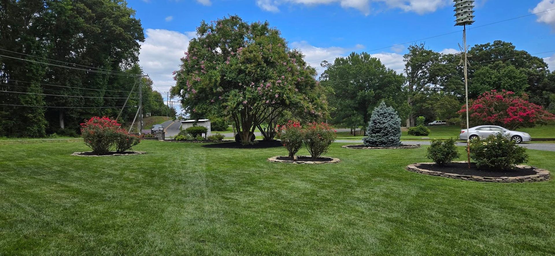 Landscaped park with green grass, flowering shrubs, trees, and a parked car under a blue sky