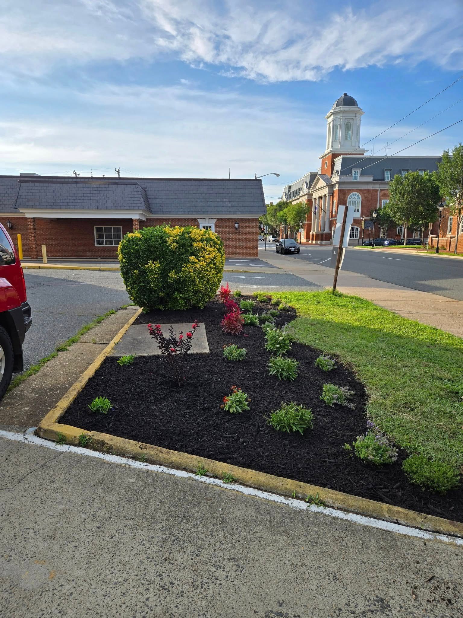 Landscaped flower bed with red blooms beside a brick building and church tower under a blue sky