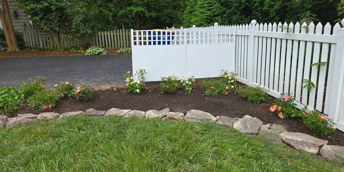 White picket fence and gate beside a curved garden bed with rocks, flowers, and green lawn in a backyard.