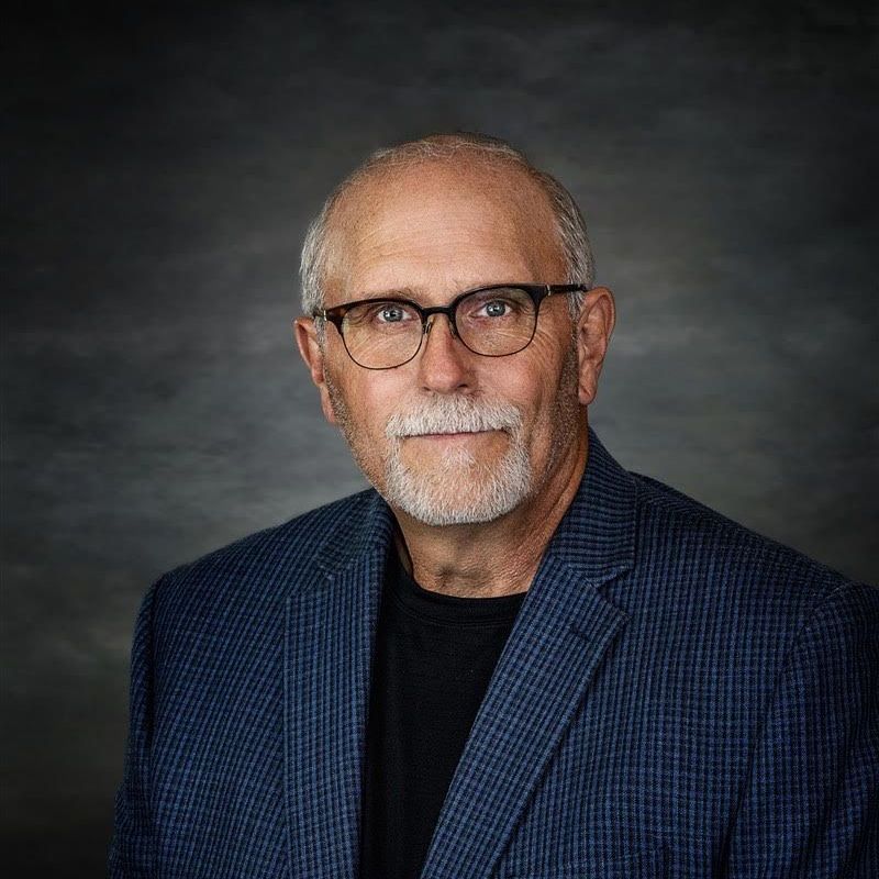 A man with glasses and a beard is wearing a blue jacket and black shirt.