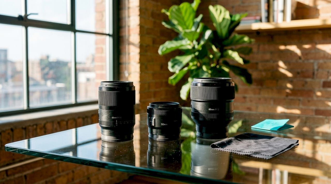 Camera lenses on a glass table in a sunlit loft with large windows and plants