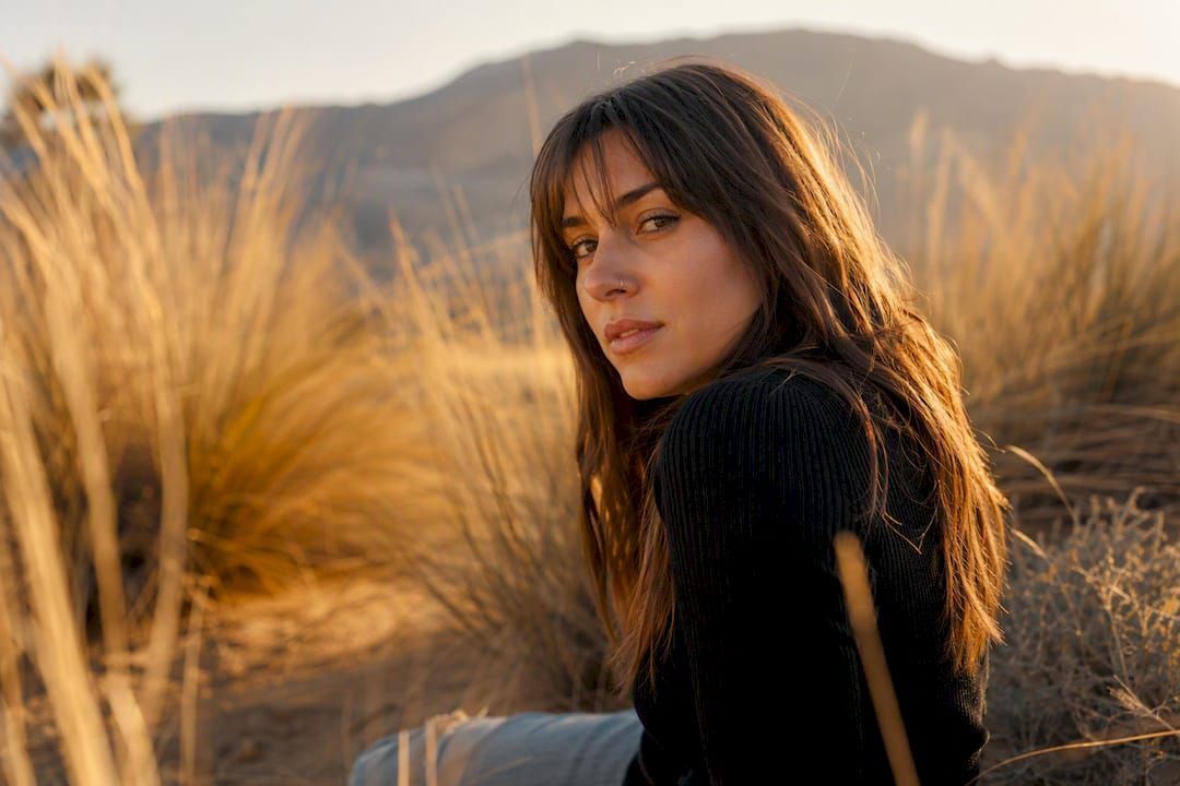 Woman in a black sweater seated in dry grass, looking back with mountains in the background.