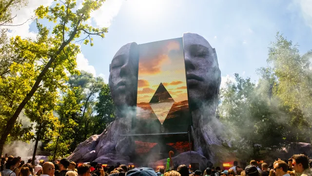 Crowd gathered before a giant stone-face stage with a glowing geometric screen outdoors in a forest setting