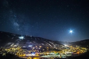 Snowy mountain town at night under a starry sky, with city lights glowing below and a bright star overhead