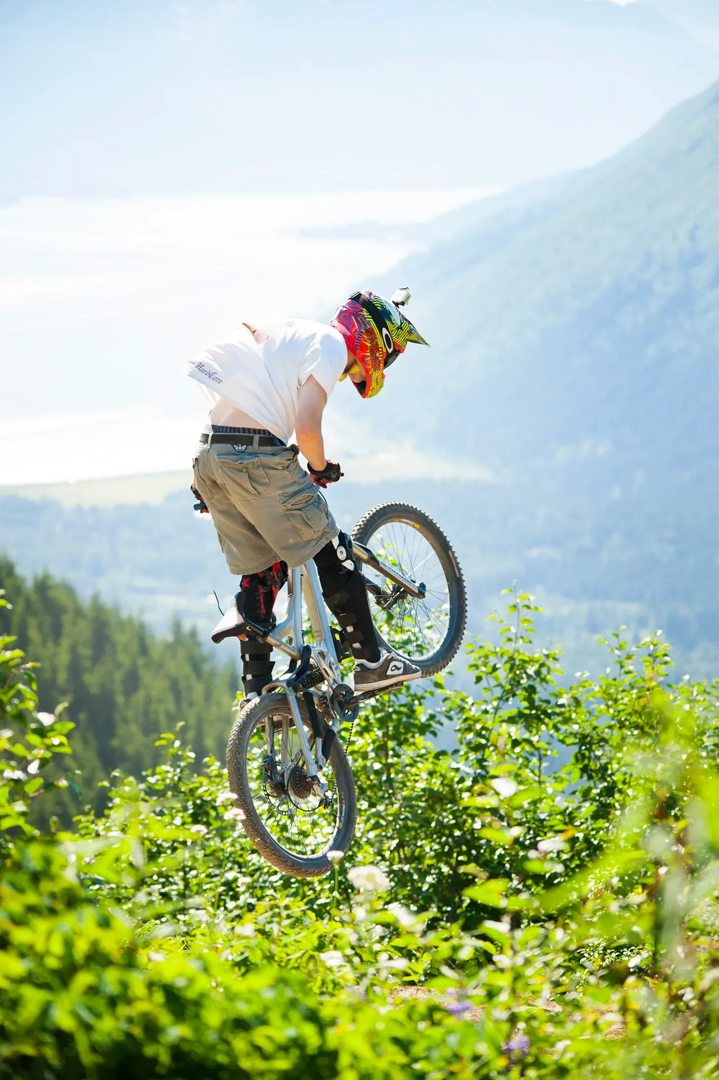 Mountain biker in a colorful helmet jumps a bike over green foliage with misty mountains behind.