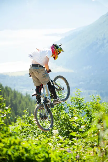 Mountain biker in a colorful helmet jumps a bike over green foliage with misty mountains behind.