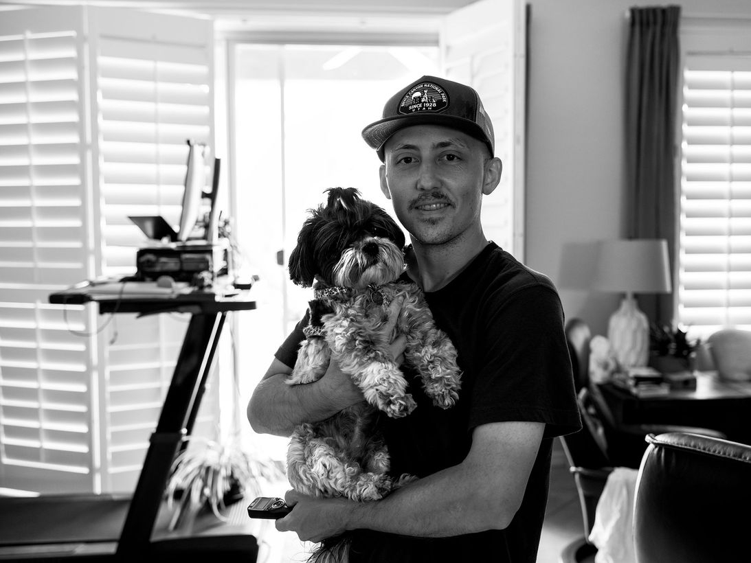 Man holding a small dog in a bright room with blinds, desk, and lamp in the background
