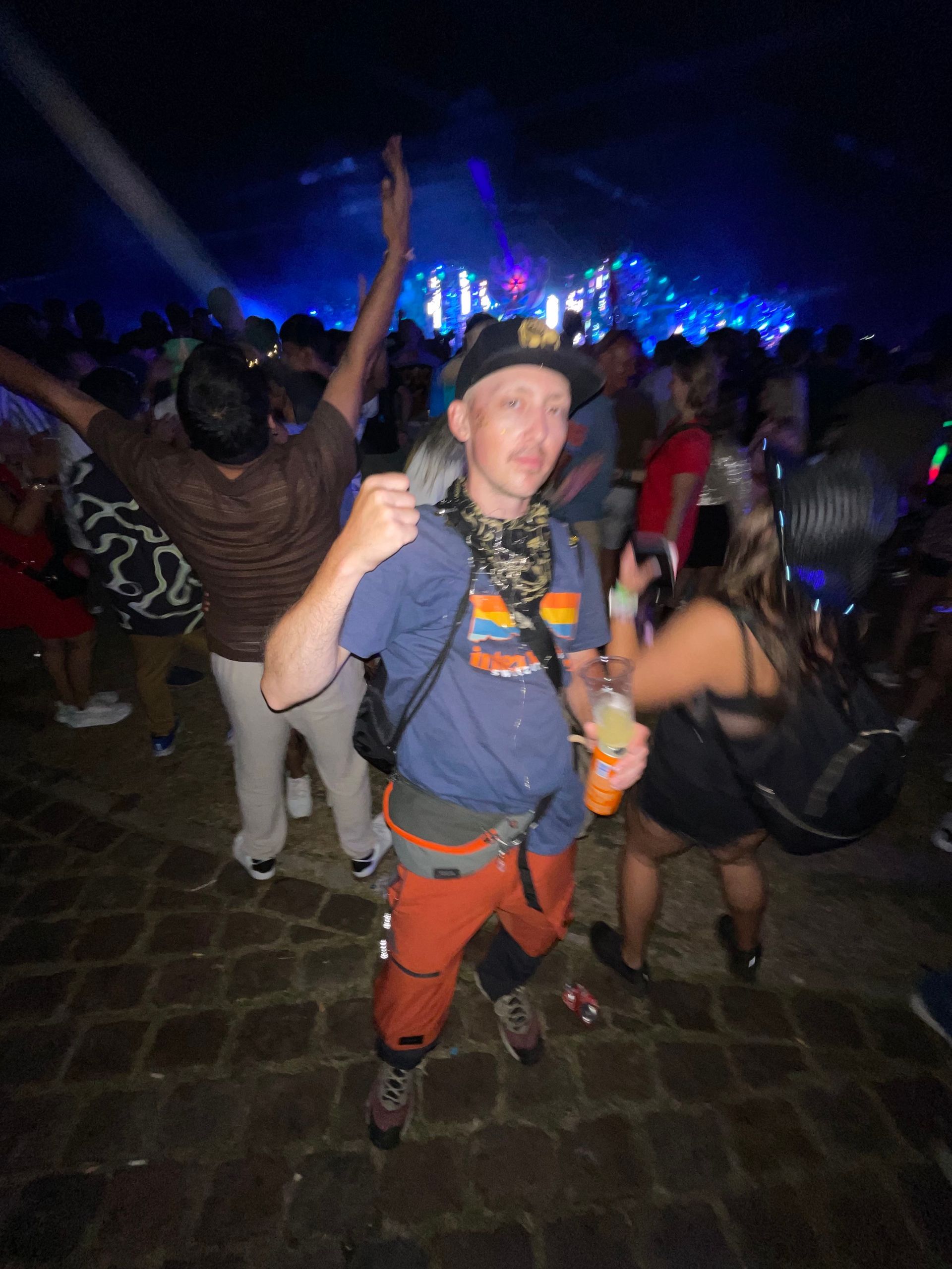 Festival crowd at night, with a person in orange pants smiling and giving a thumbs-up near stage lights.