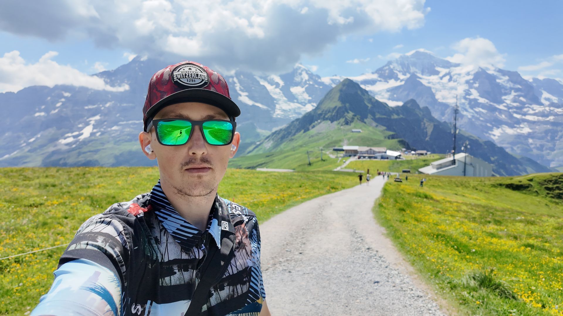 Selfie on a mountain trail with snow-capped peaks, green slopes, and a cloudy blue sky.