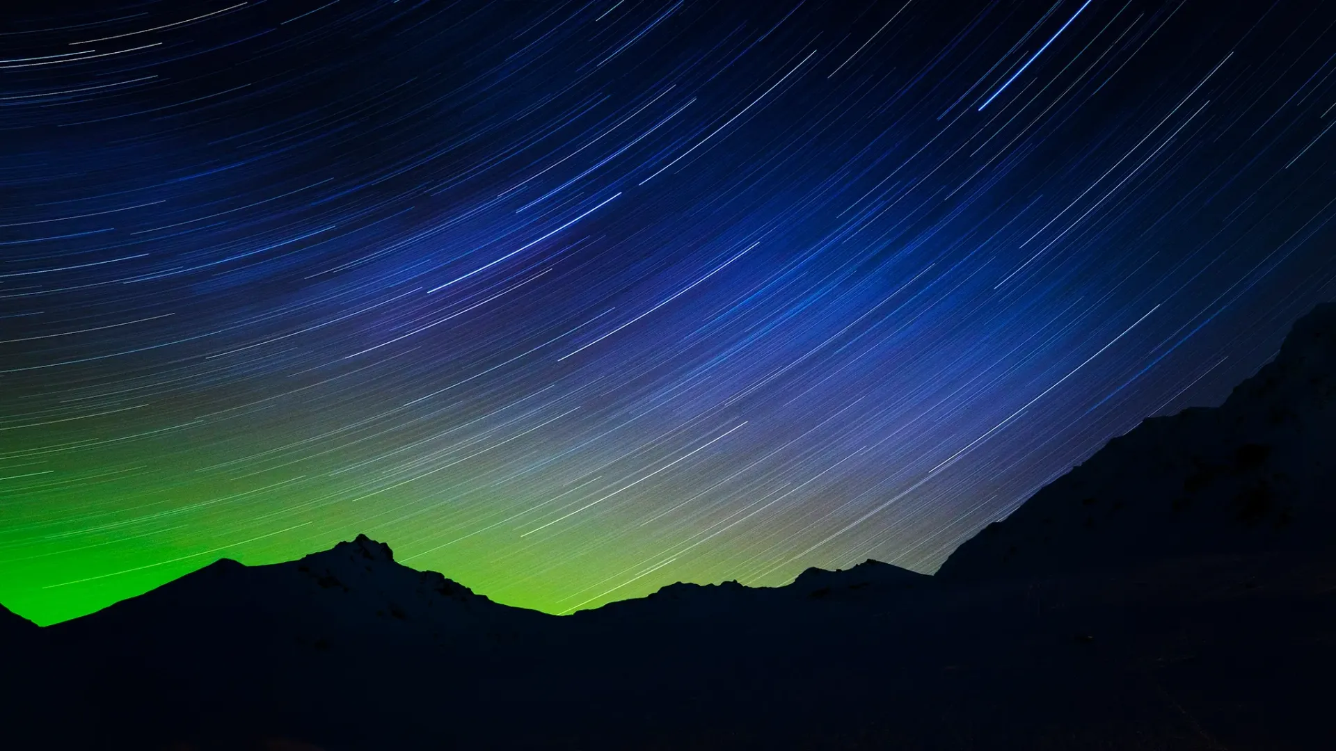 Star trails over dark mountain silhouettes with a green glow on the horizon