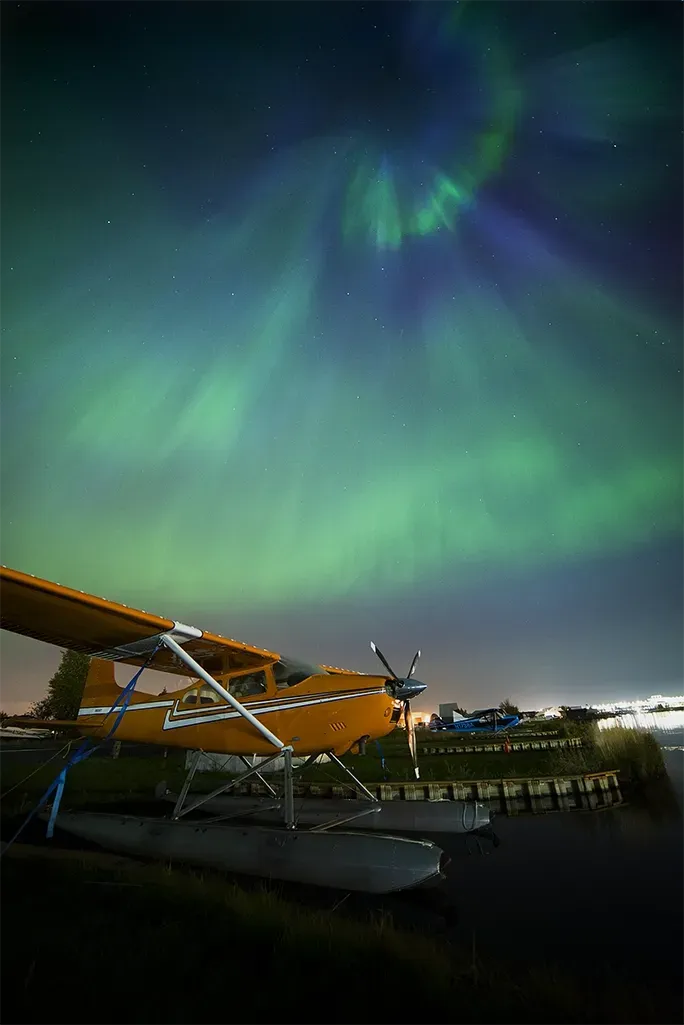 Aurora borealis glowing green and blue over a waterfront with a docked seaplane at night