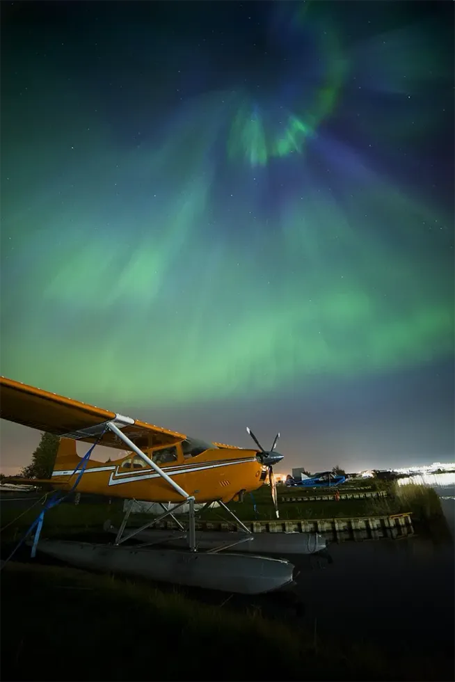 Aurora borealis glowing green and blue over a waterfront with a docked seaplane at night