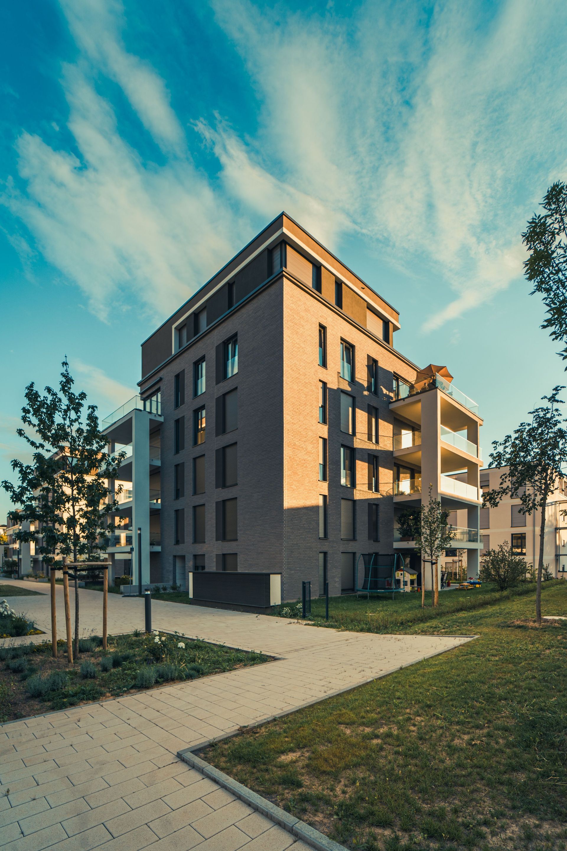 Modern apartment building at sunset with a landscaped path, trees, and blue sky
