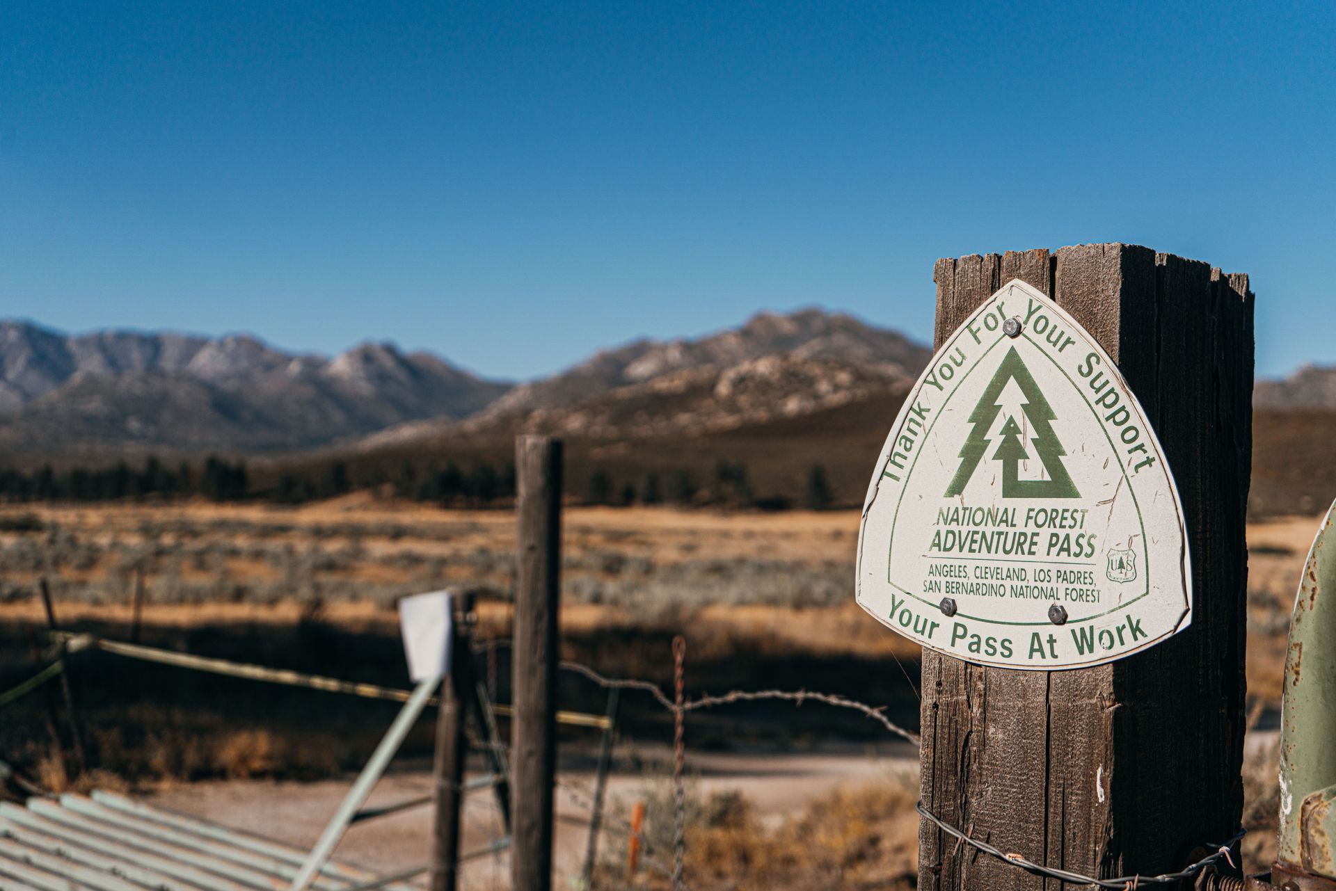 Trail sign on a wooden post in a fenced field with mountains under a clear blue sky.
