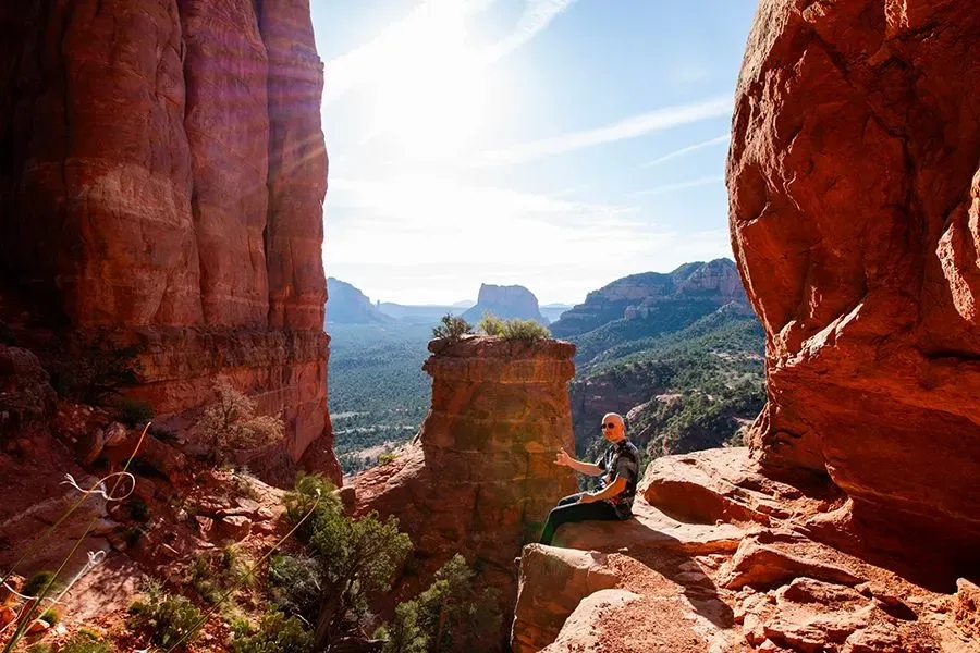 Sunlit canyon overlook with red rock cliffs and a person sitting on a ledge, viewing distant mesas