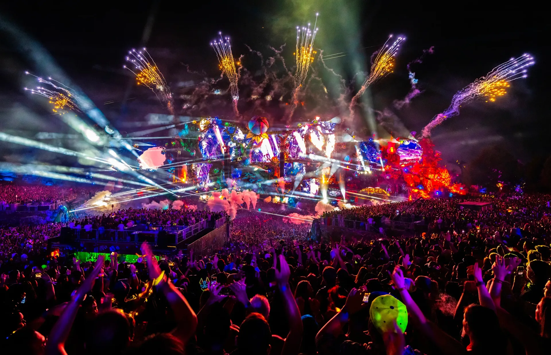 Crowded outdoor concert with colorful stage lights, fireworks, and cheering crowd with raised hands