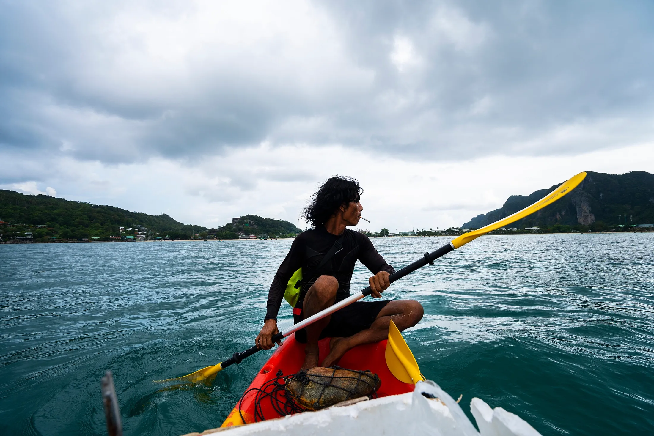 A man paddles a red kayak across calm teal water, sitting cross-legged with a paddle mid-stroke and a hand-rolled cigarette between his lips. He wears a black rash guard and yellow vest, with dark tousled hair.