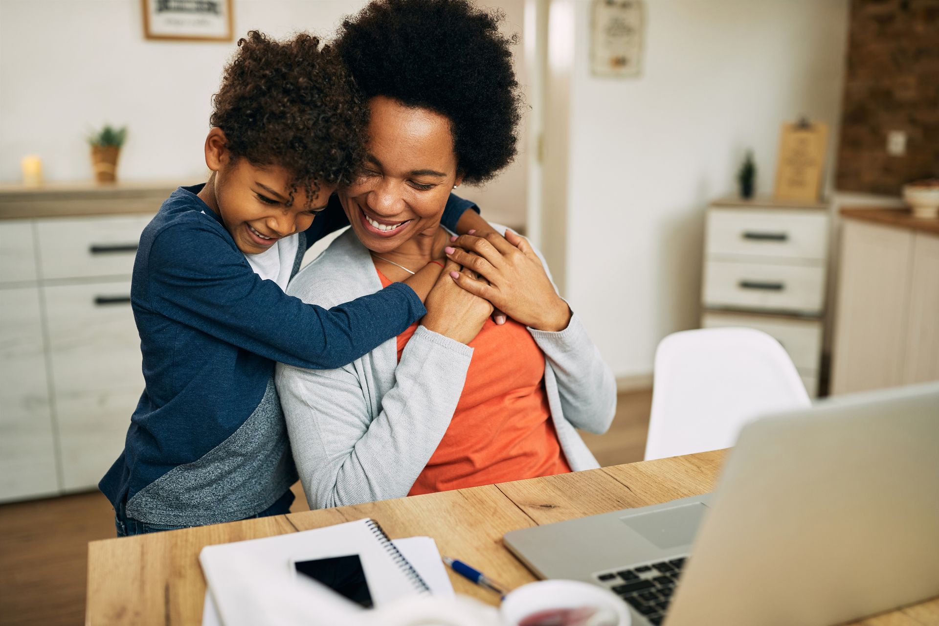 A woman and a child are hugging while sitting at a table with a laptop.