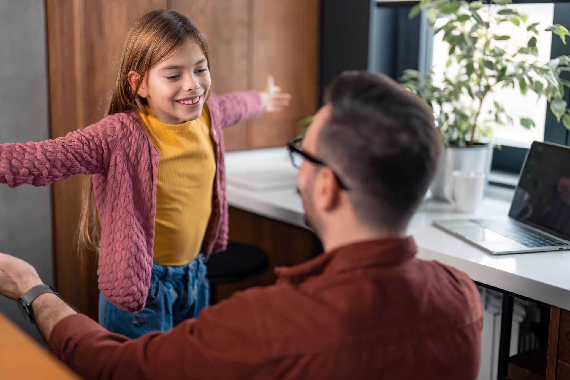 A man is sitting at a desk with a laptop and a little girl is standing next to him.