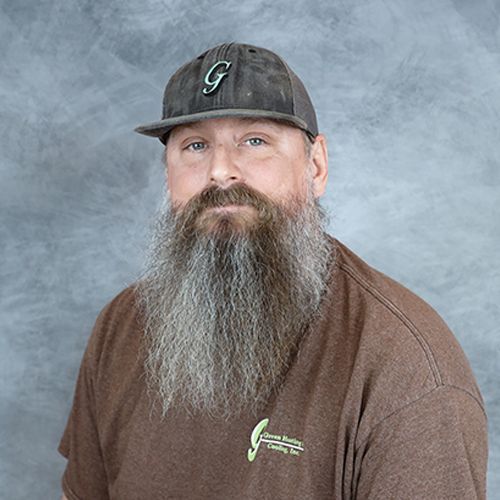 Man with long gray beard, wearing a baseball cap and brown shirt with logo on gray background.