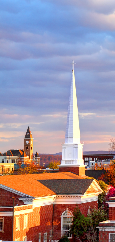 Town skyline with red-brick rooftops and a tall white church spire under a cloudy sunset sky