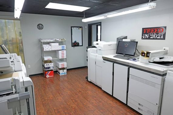 Office with printers, desks, and shelves in a gray room with wood flooring.
