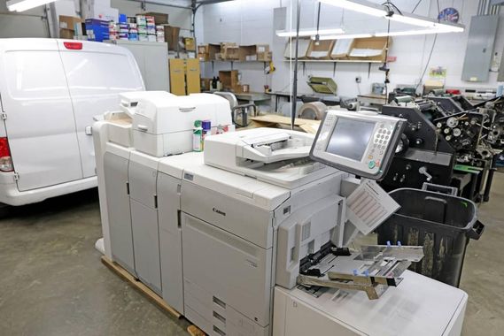 Industrial office with large white printers/copiers, a van, and warehouse shelves in the background