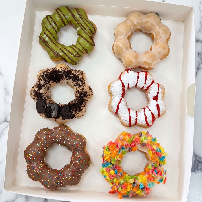 Six assorted decorated donuts in a white box, topped with sprinkles, glaze, and icing.