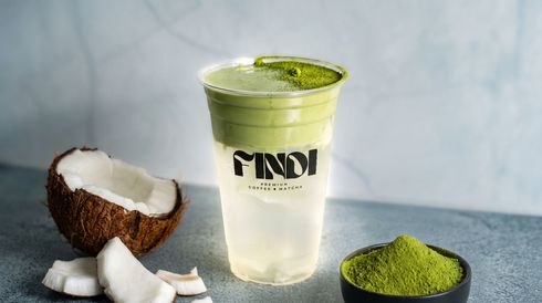 Clear cup of coconut drink with green powder on top, beside coconut pieces and a bowl of matcha powder.