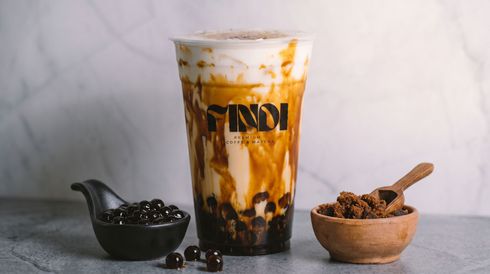Iced coffee in a clear cup with caramel drizzle, flanked by coffee beans in small bowls on a gray surface.