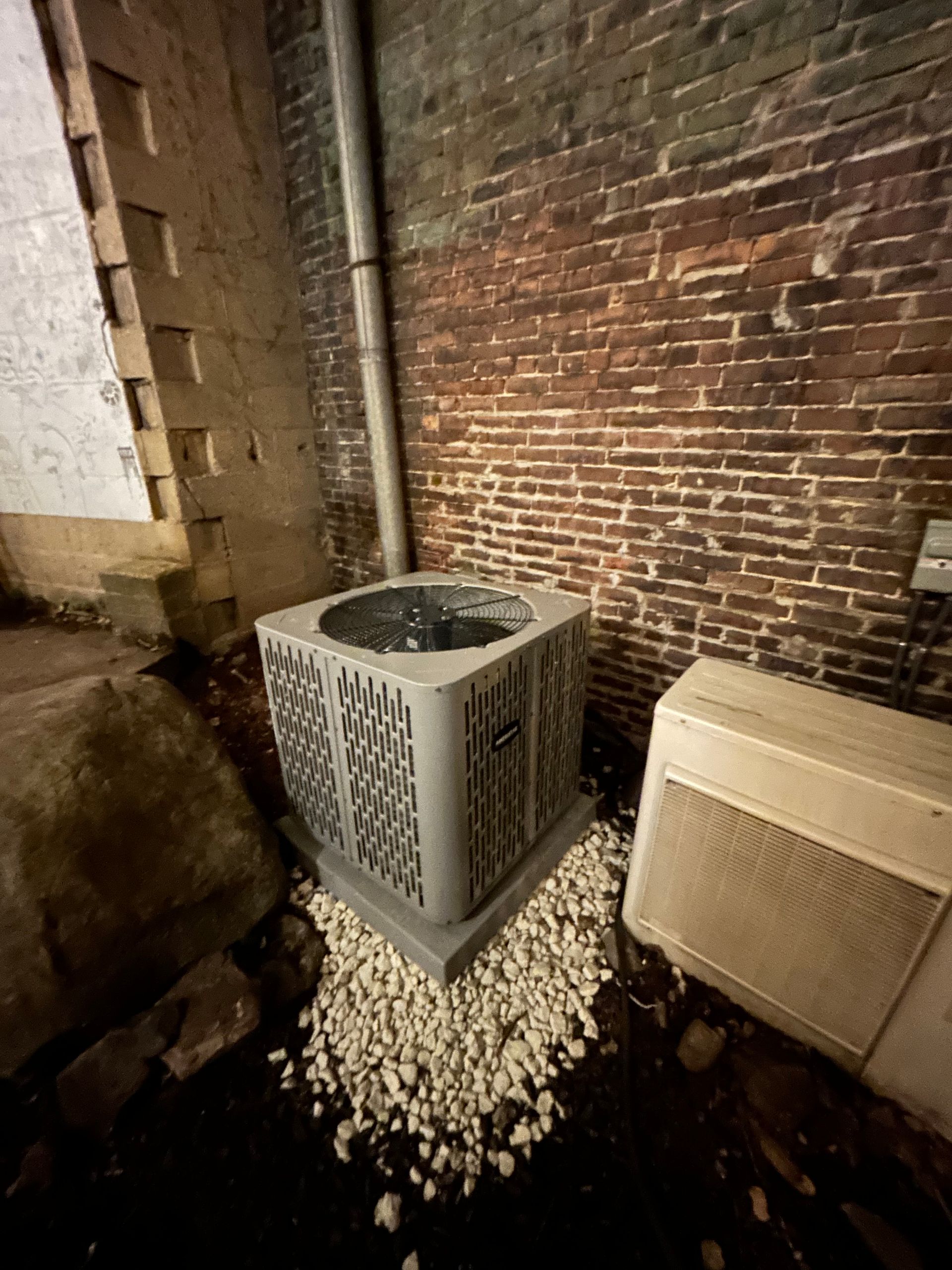 An outdoor air conditioning unit next to a brick wall, on a bed of small rocks.