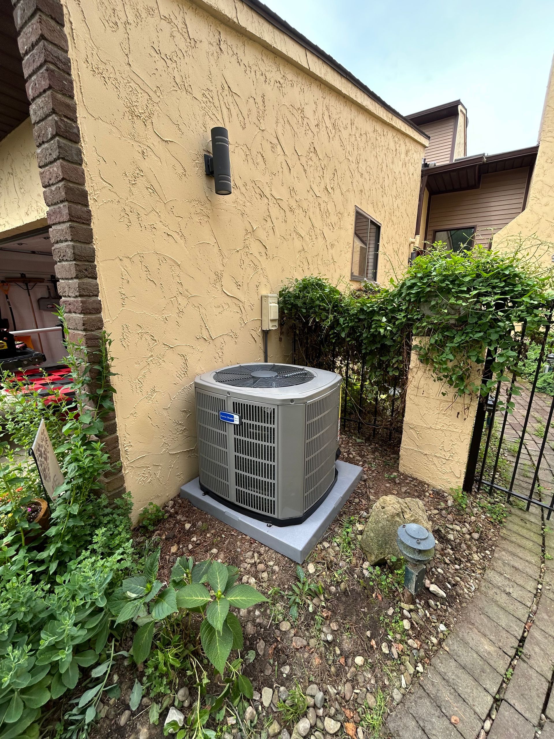 Air conditioning unit on a concrete pad next to a building with brown stucco. Shrubs and ground cover surround it.