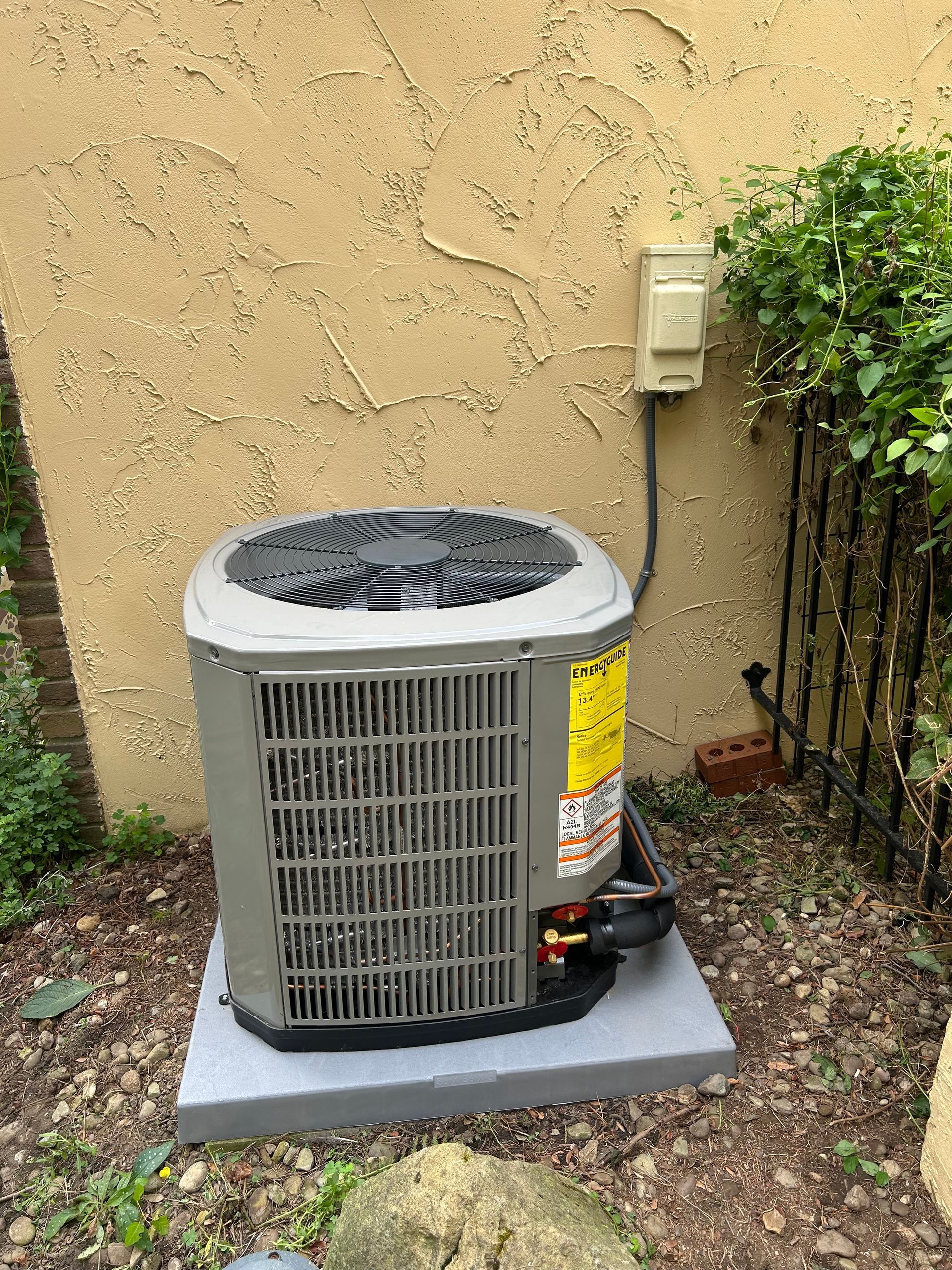 Outdoor air conditioning unit on a concrete pad, near a stucco wall and bushes.