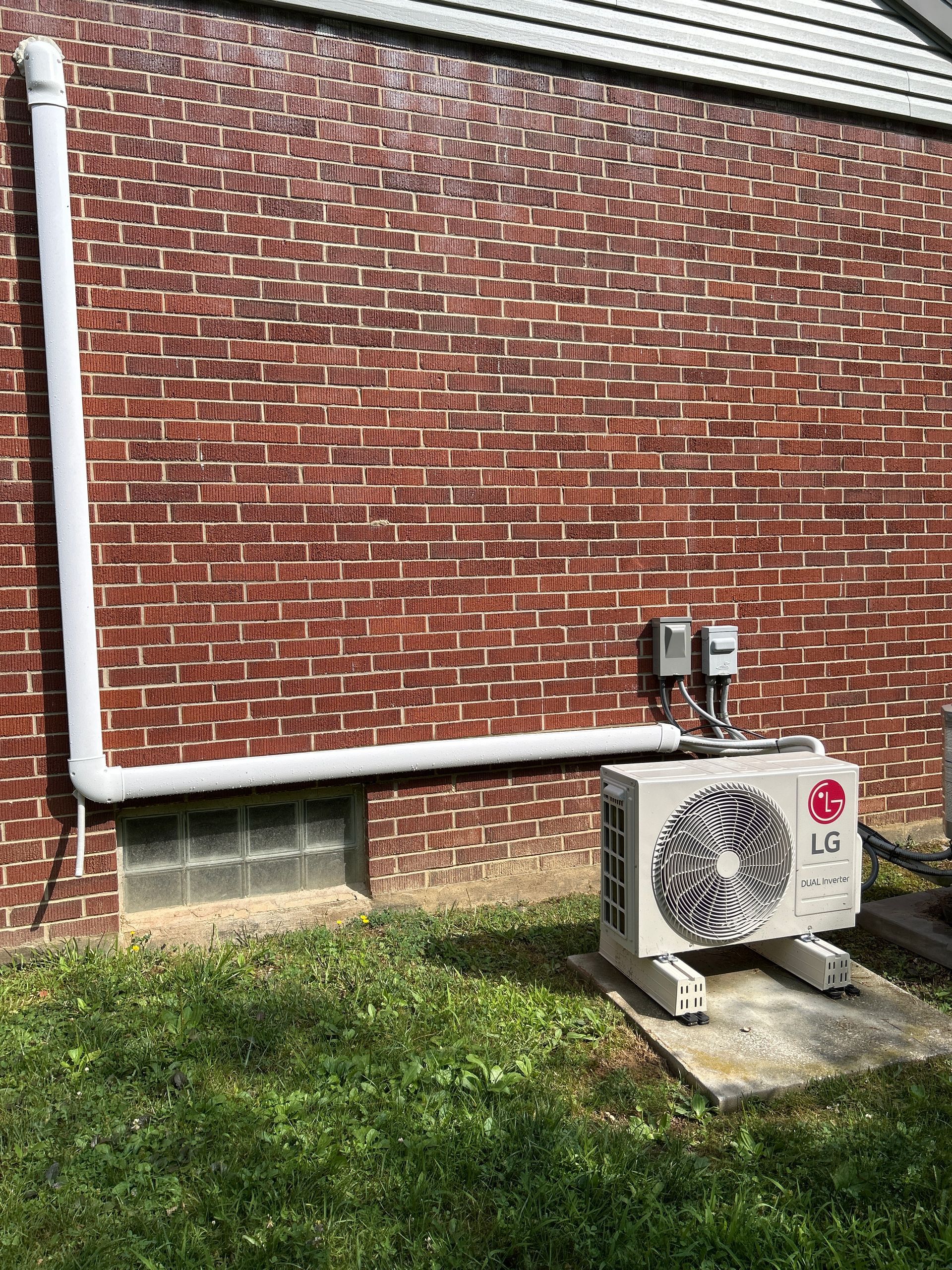 LG air conditioner unit against a red brick wall with white pipes; set in a grassy yard.