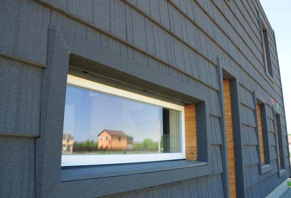 Blue-gray building exterior with a large rectangular window reflecting a small house and sky.