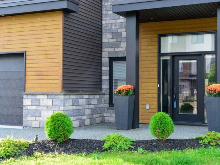 Modern house exterior with gray stone, wood siding, black front door, and small landscaped shrubs
