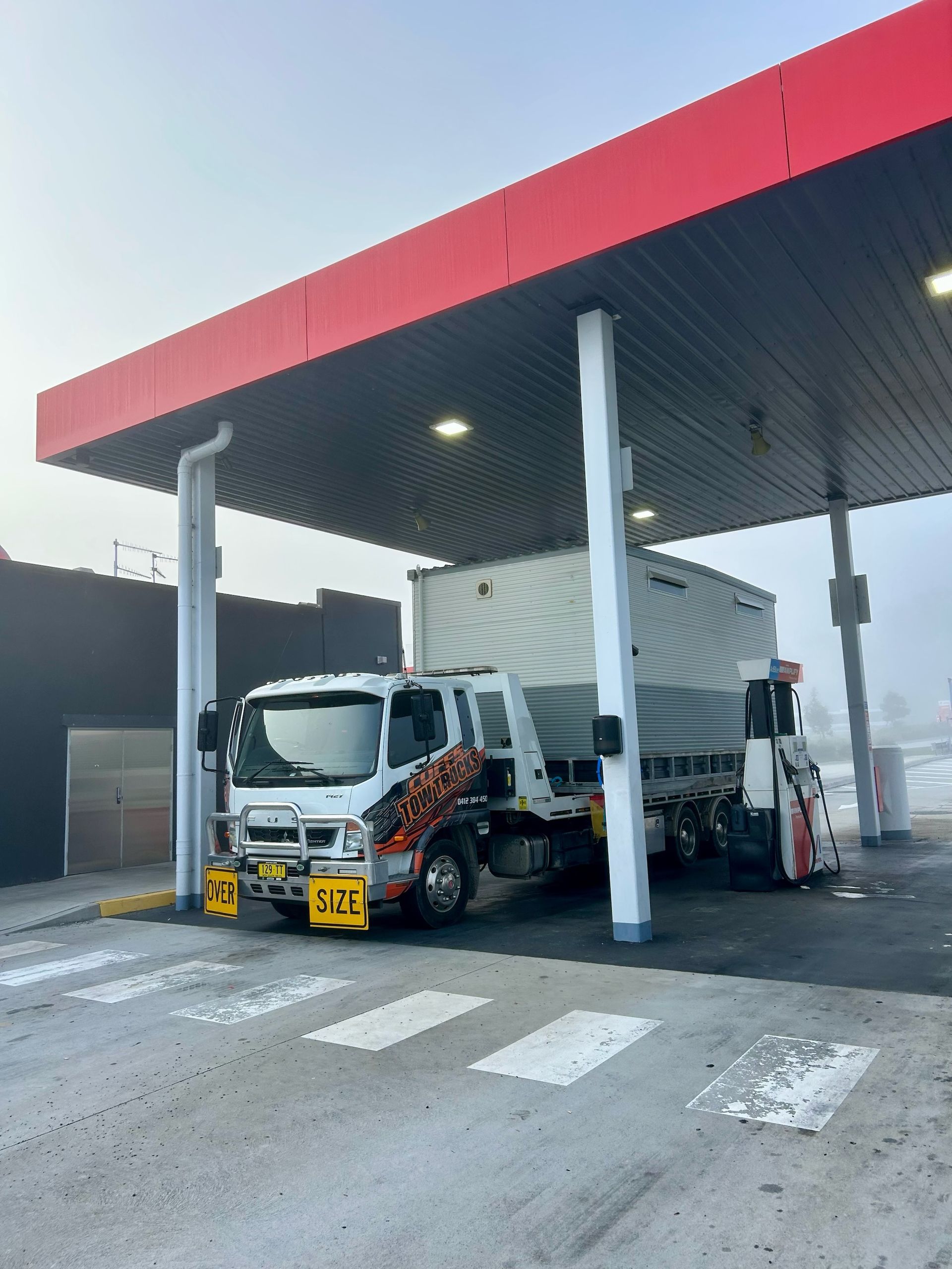 Fuel Truck Parked Under A Red Gas Station Canopy In A Rainy Lot — Coffs Tow Trucks In Woolgoolga, NSW