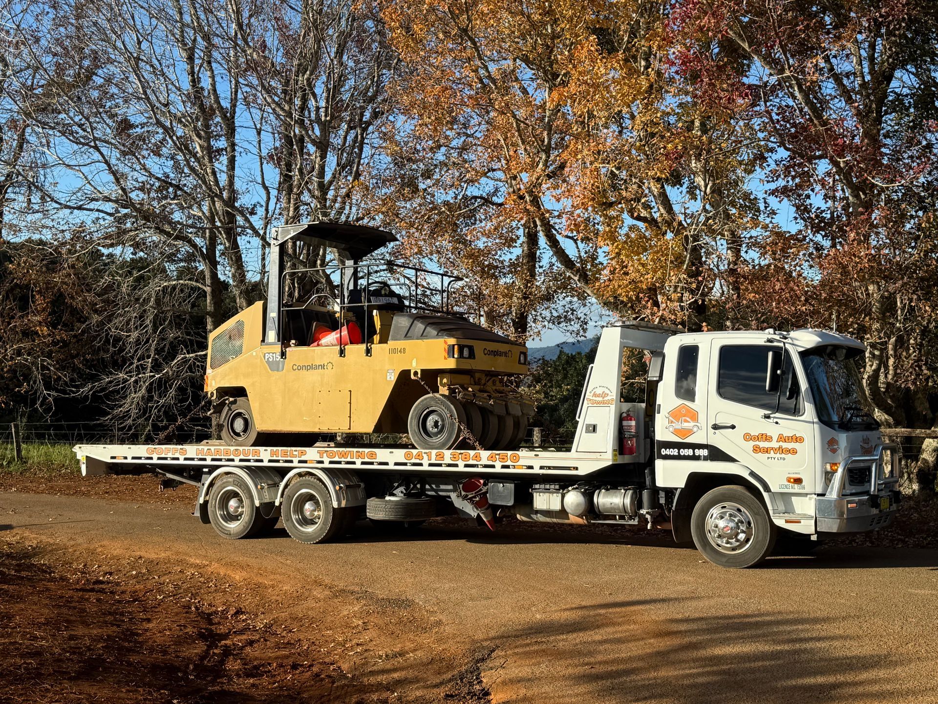 Tow Truck Hauling A Yellow Forklift On A Flatbed In A Wooded Area During Fall — Coffs Tow Trucks In Woolgoolga, NSW