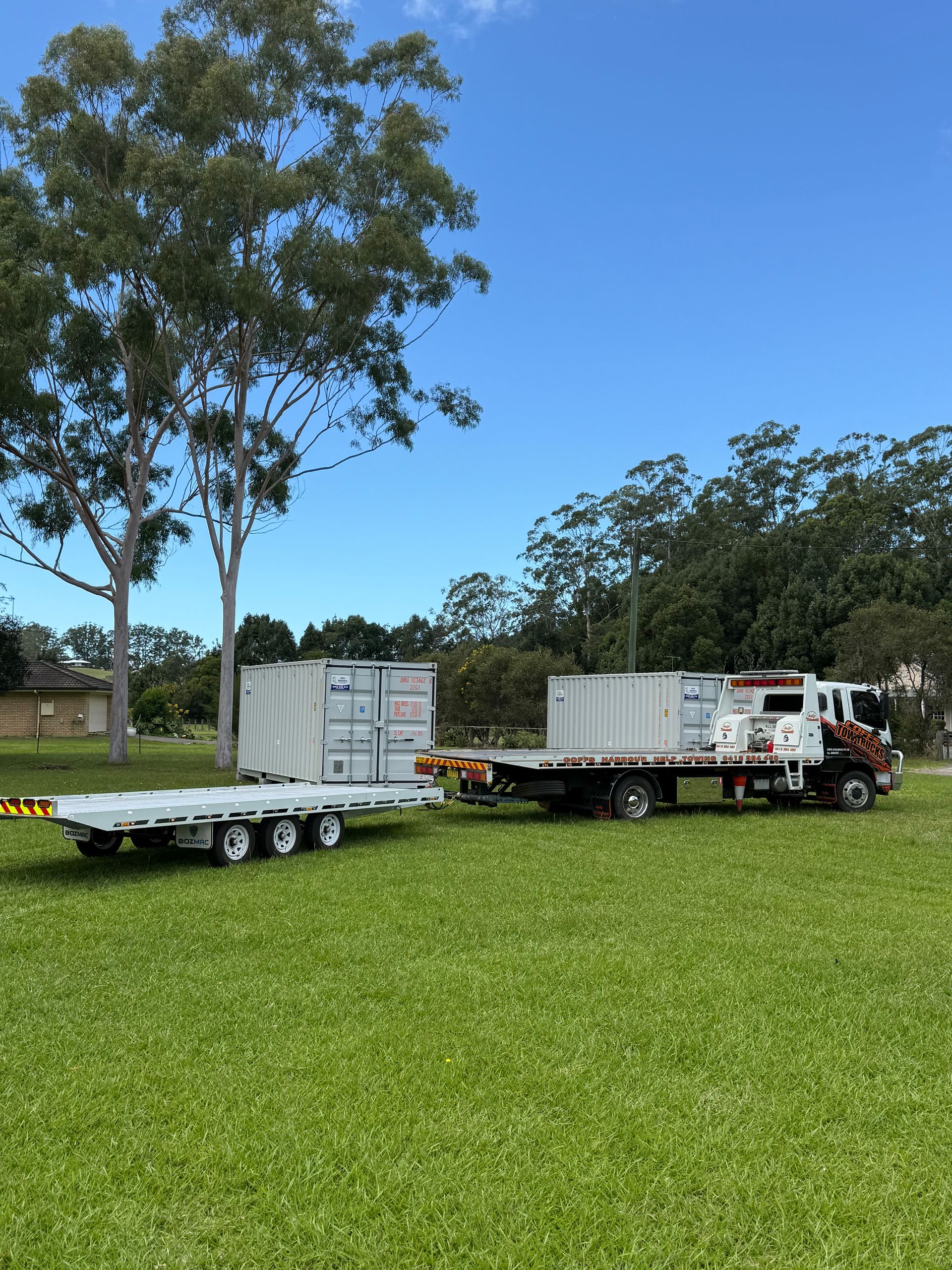 Flatbed Trucks And Trailers Parked On Grass Under Tall Trees — Coffs Tow Trucks In North Boambee Valley, NSW