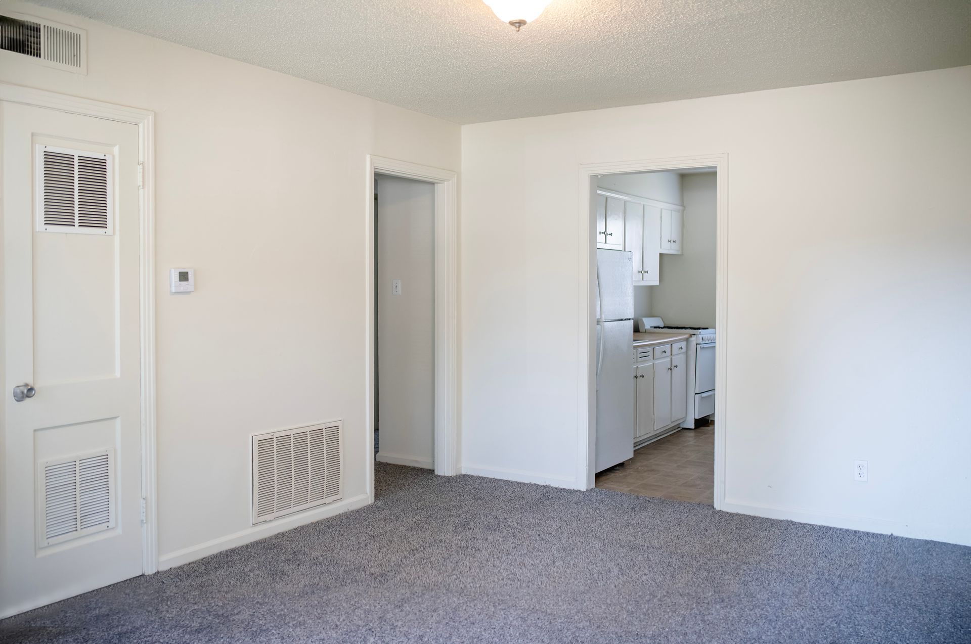 An empty living room with a gray carpet and white walls leading to a kitchen.