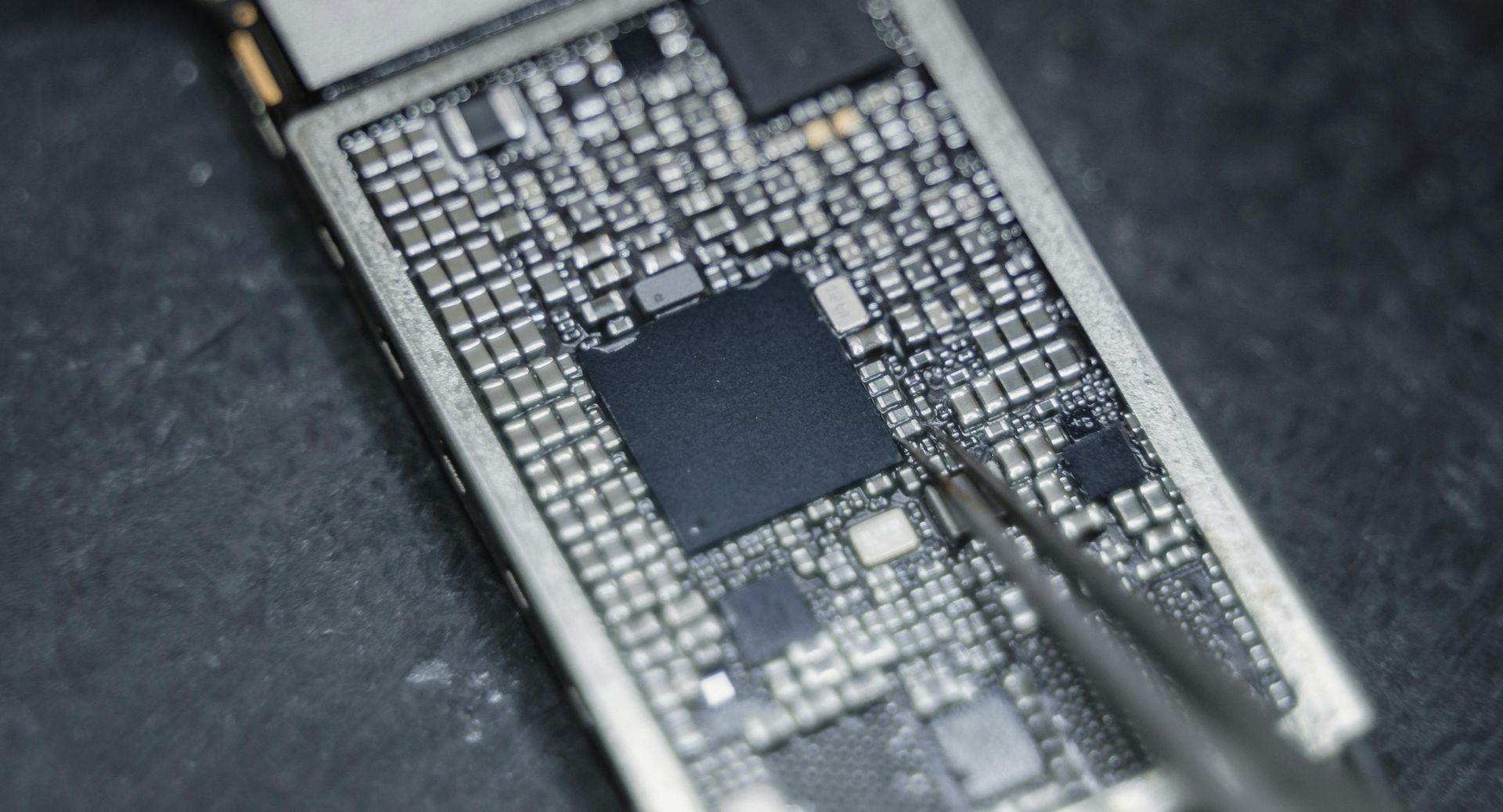 Close-up of a circuit board with many tiny electronic components, being handled with tweezers.