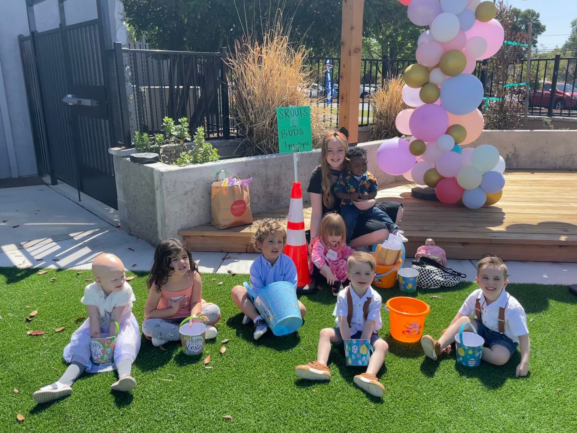 A group of children are sitting on the grass with buckets and balloons.