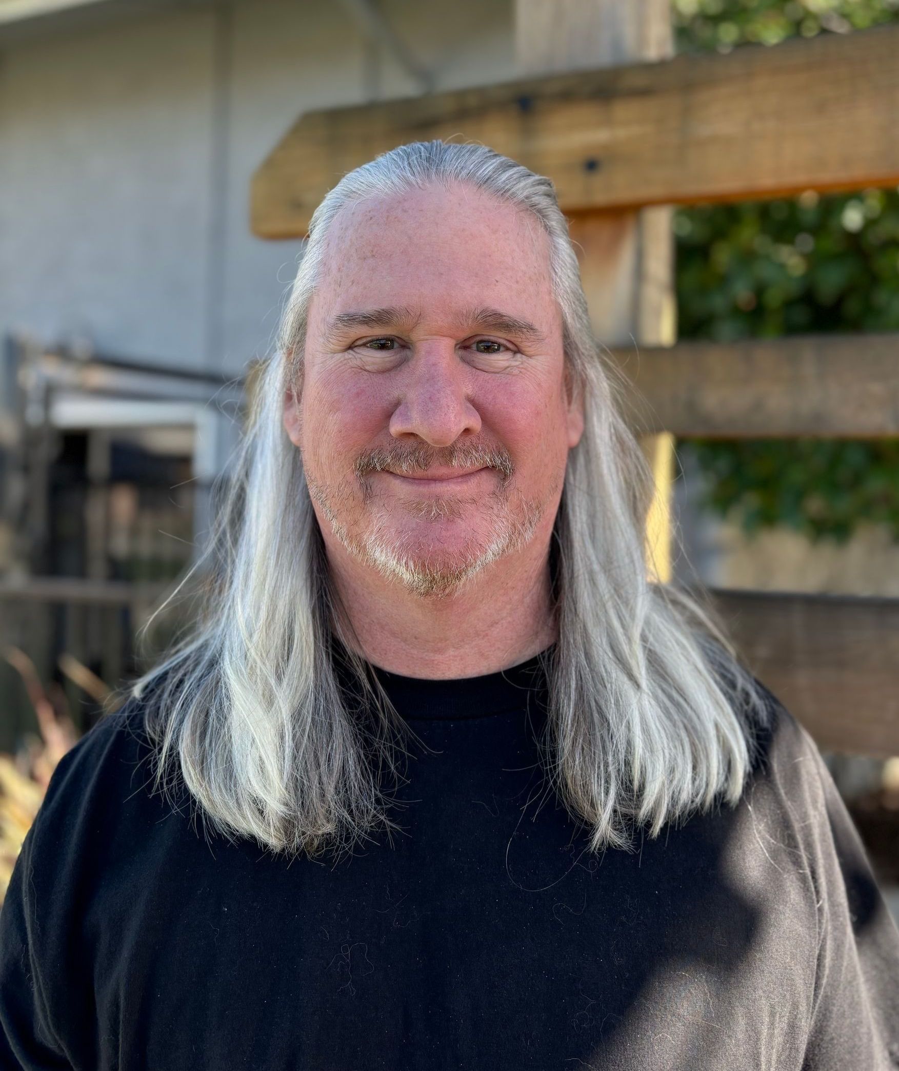 A man with long white hair and a beard is wearing a black shirt and smiling.